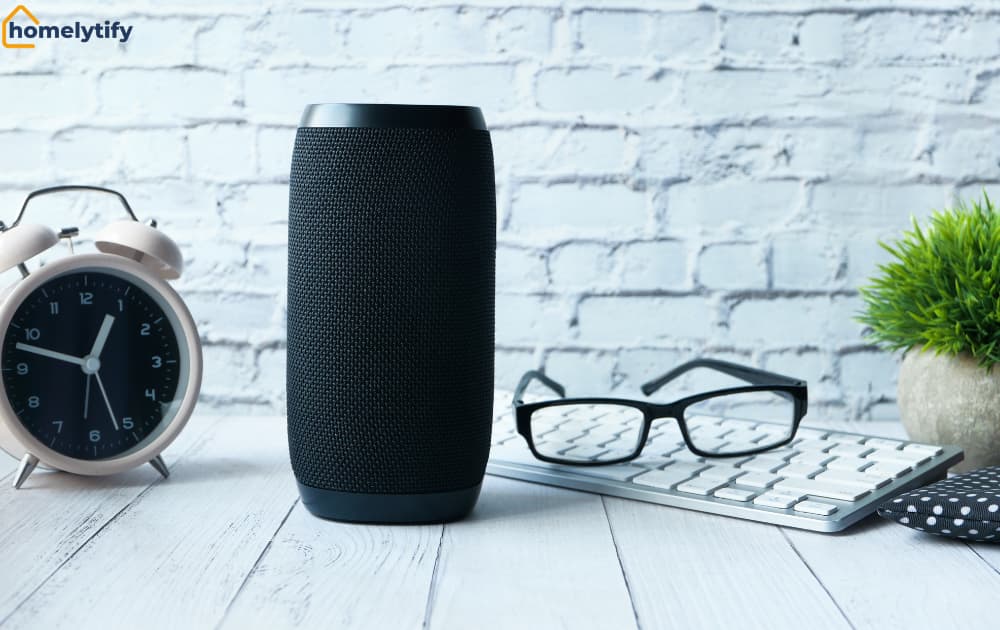 Black smart speaker on a desk beside a clock, keyboard, and glasses.