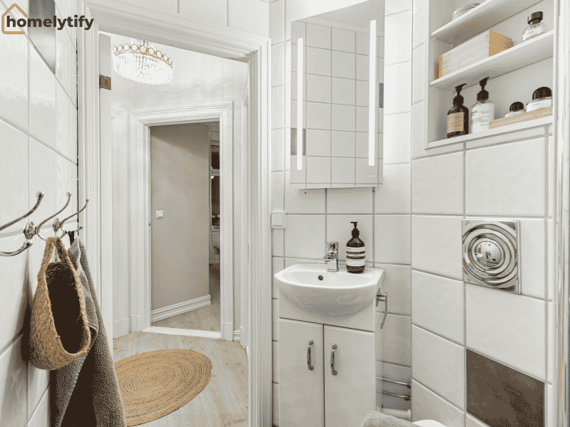 White tiled bathroom with wall-mounted shelves, under-sink cabinet, and hooks for bags and towels, showing smart bathroom small space storage ideas