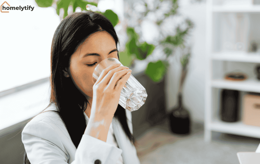 Woman drinking water from a clear glass in a bright indoor space with green plants in the background.