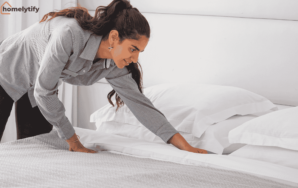 Woman making her bed by straightening white sheets and pillows in a clean bright bedroom.