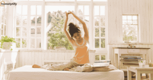 A woman stretching on a bed near large windows as part of a morning routine for beginners in a calm and sunlit bedroom