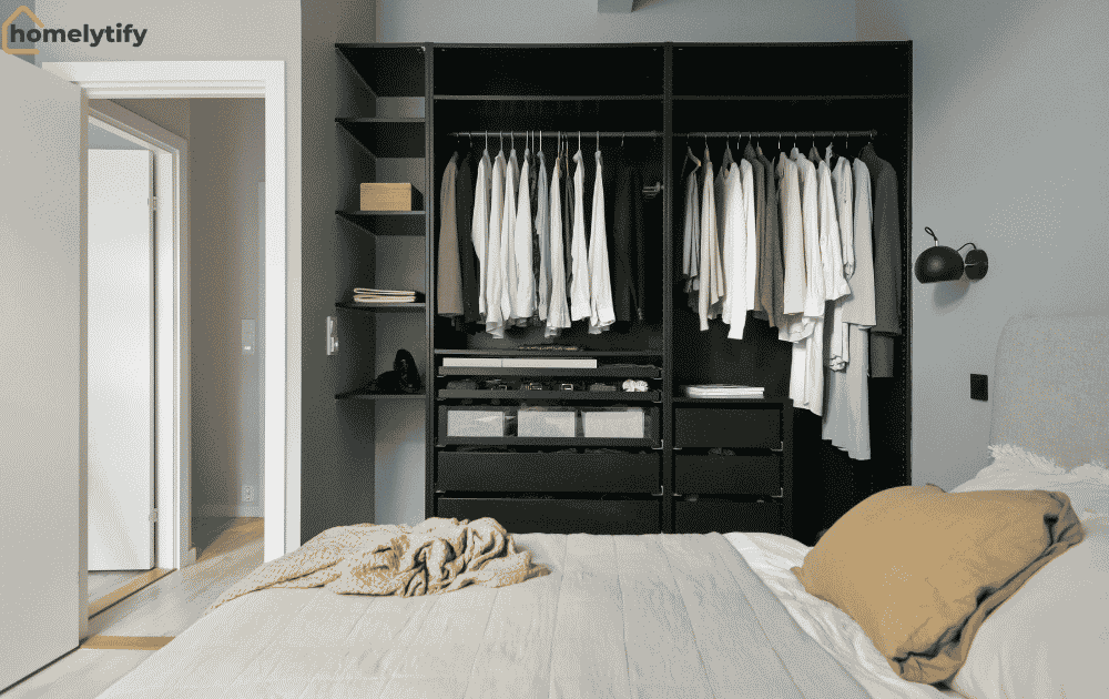 Organize a small bedroom on a budget with a dark open wardrobe system featuring hanging clothes, built-in drawers, and open shelves beside a neatly made bed with neutral pillows in a modern minimalist bedroom.