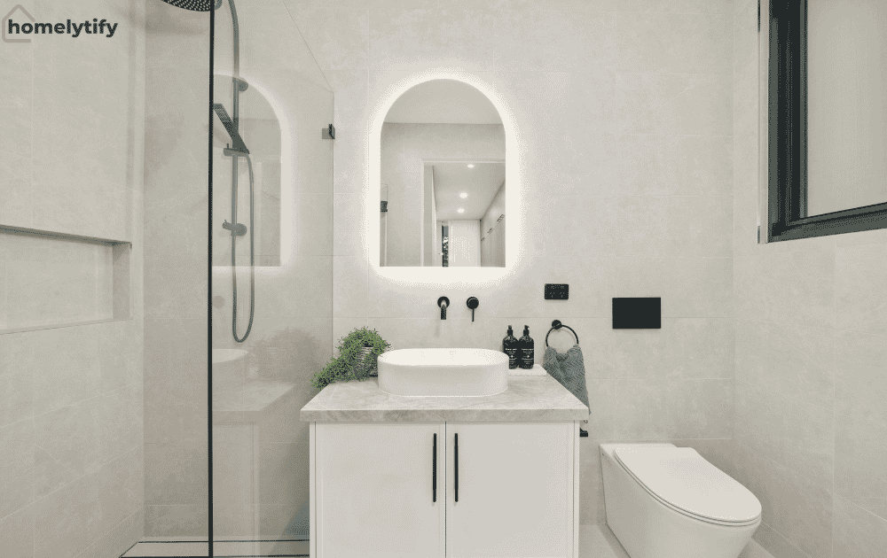 Simple small bathroom organization hacks with a white under-sink cabinet, LED arch mirror, vessel sink, glass shower enclosure, and clutter-free countertop for a clean, modern small bathroom setup.