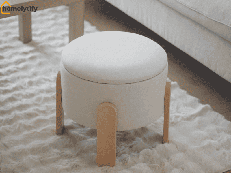 A white round storage ottoman with wooden legs placed on a fluffy rug beside a bed, perfect for organizing a rental home