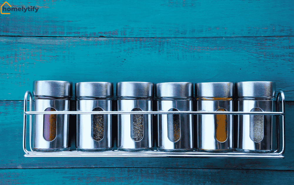 Wall-mounted spice rack holding six stainless steel spice jars neatly organized against a rustic teal wooden background.