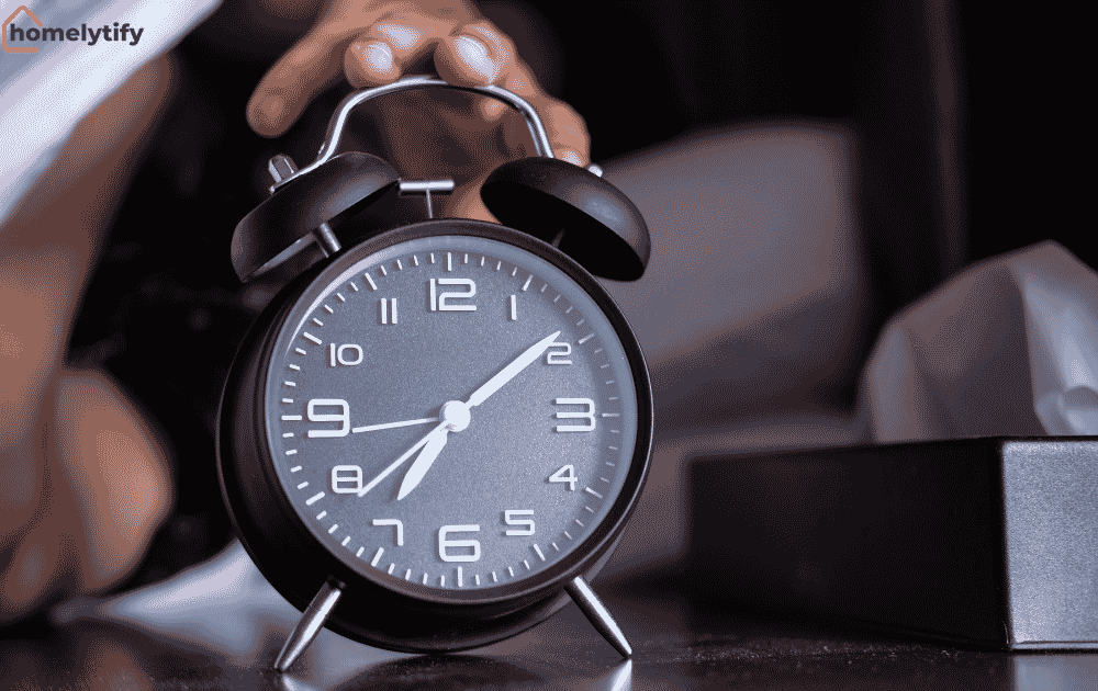 A hand reaching for a black alarm clock in bed to build 7 evening routine habits for busy people.