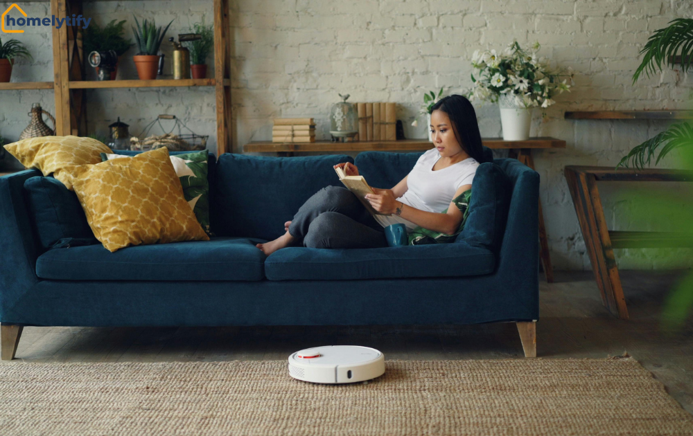 Woman reading a book on a blue sofa while a robot vacuum runs — relaxing evening habits for a calmer home