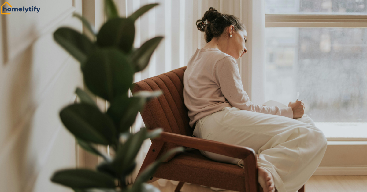 A woman practicing relaxing evening habits by journaling quietly in a cozy armchair by the window with an indoor plant nearby 