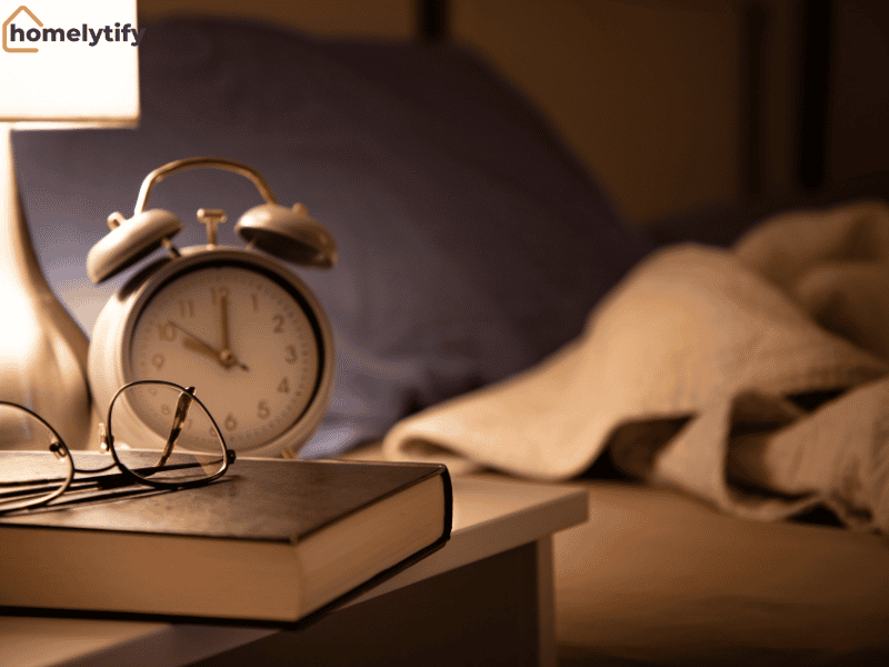 An alarm clock and glasses on a book beside a cozy bed at night, reminding you to set a sleep anchor time for better rest